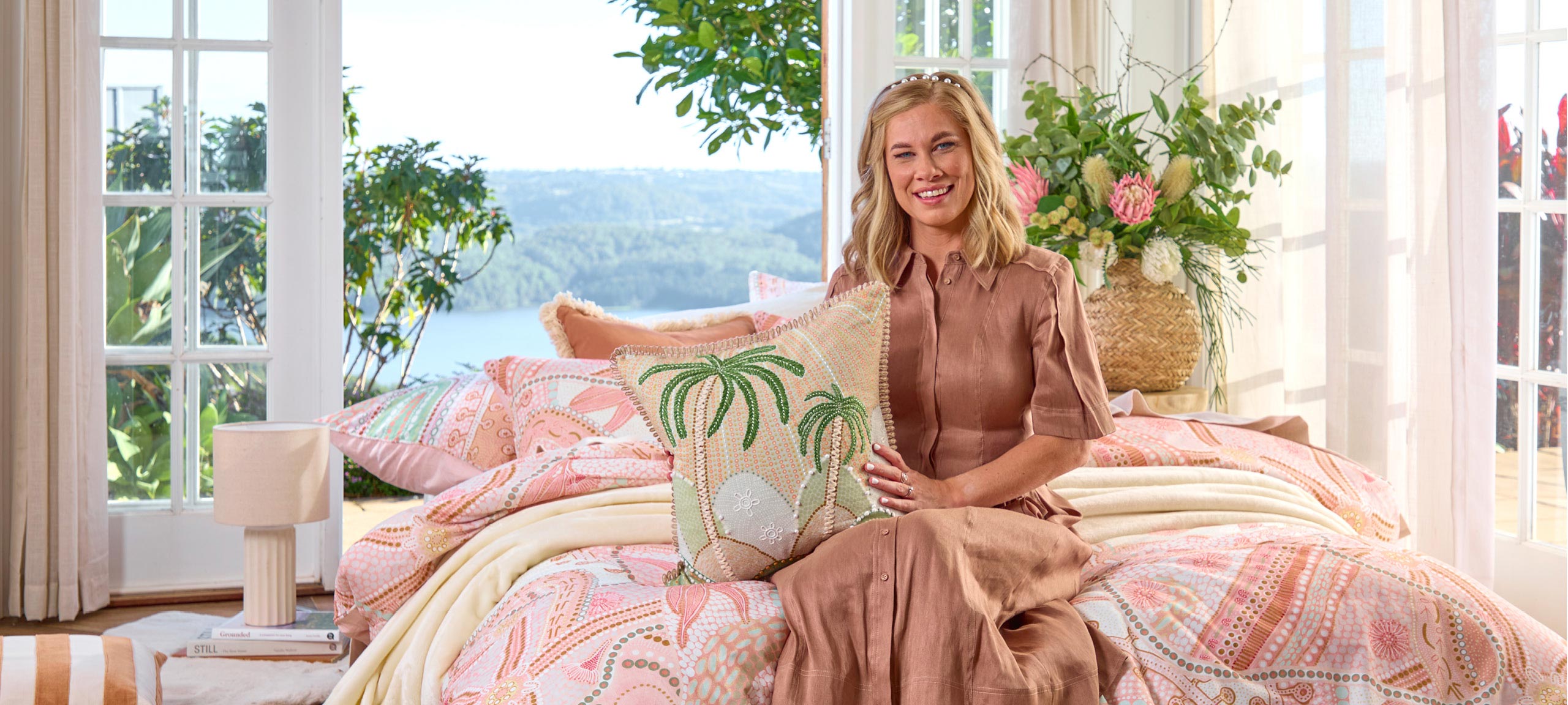 The picture features Domica Hill, a blonde woman, holding a cushion from the Coastal Connections collection that features a palm tree design in an Australian Indigenous art style. Domica is sitting on a bed that features a quilt cover set from the Native Country collection featuring a bright Indigenous Australian design.