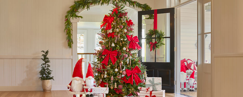 A Christmas Tree sits in the middle of a room, decorated with traditional red and white ornaments. Next to it sits two santa gnomes and a pile of presents.